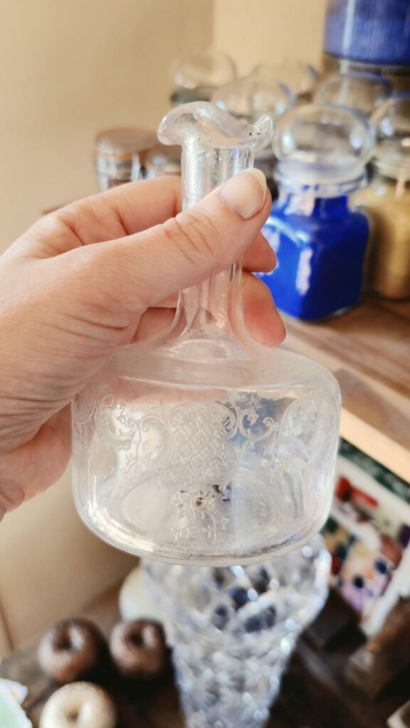 Carafe ancienne en verre gravé tenue à la main, devant des bocaux de pigments